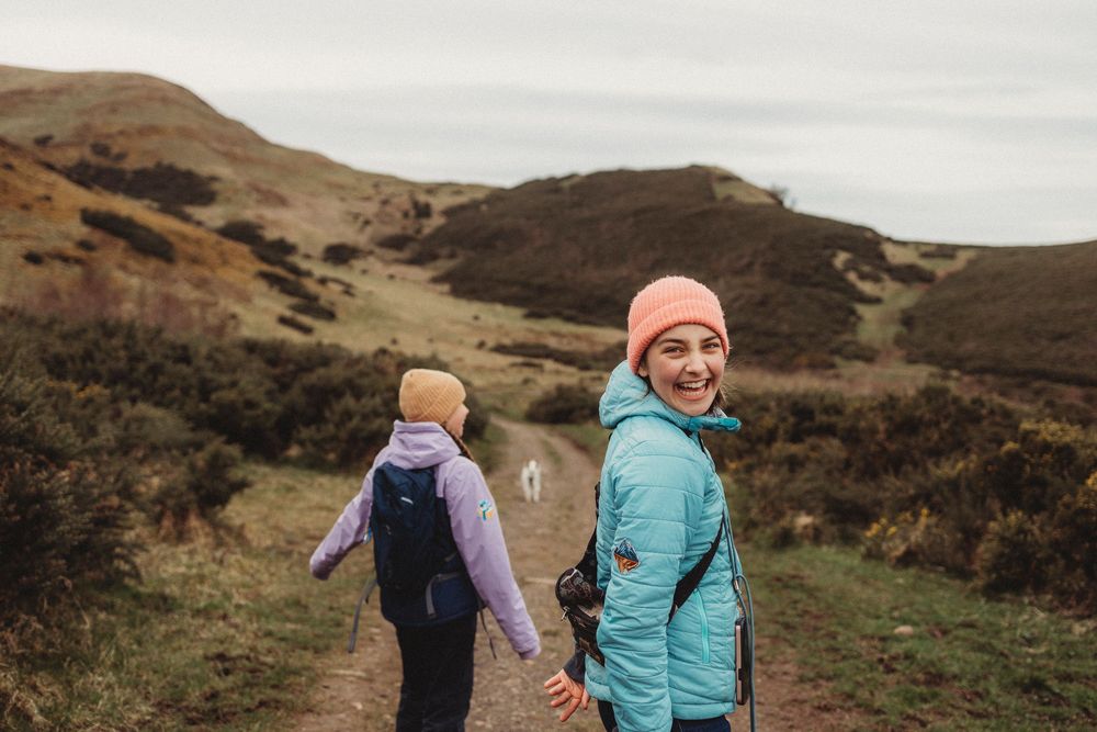 Two women hiking on a scenic trail through rolling hills, one smiling back at camera wearing a pink beanie and blue jacket.