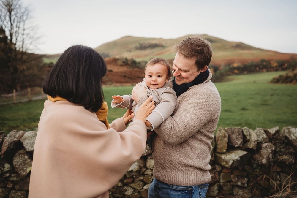 A young family with a baby enjoying an outdoor moment by a stone wall with green hills in the background.