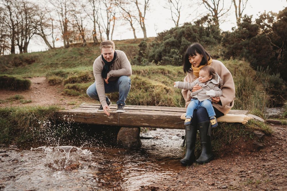 Family of three enjoying nature outdoors, parents sitting on wooden bridge over stream with baby in autumn countryside.