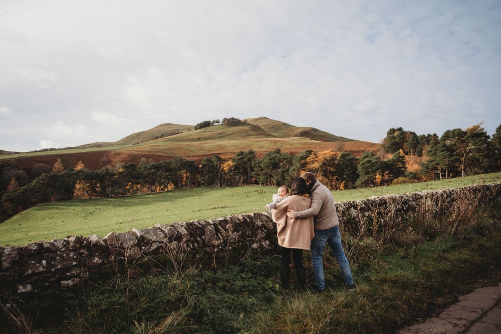 Couple embracing by a stone wall with rolling green hills and autumn foliage in a scenic countryside landscape.