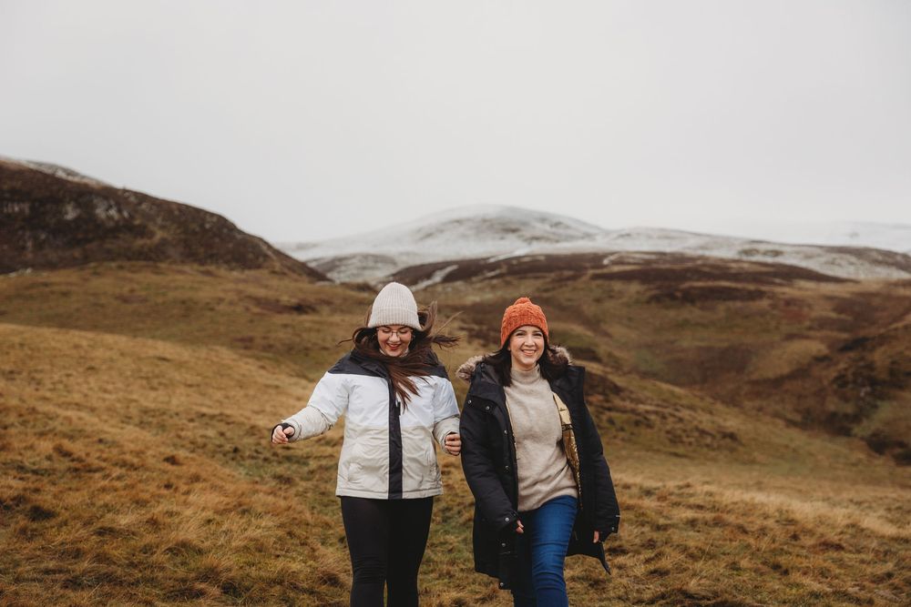Two women hiking in winter hats and jackets on a misty highland landscape with snow-capped mountains.