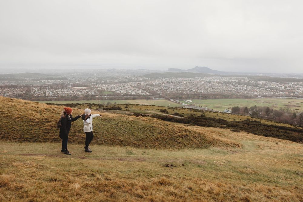 Two hikers pointing at a panoramic cityscape view from a grassy hilltop on a cloudy day.
