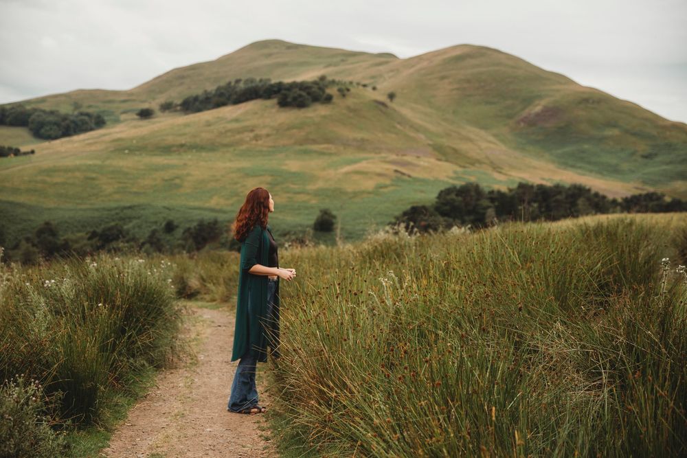 A red-haired woman in a teal dress stands on a dirt path surrounded by lush green hills and tall grass.