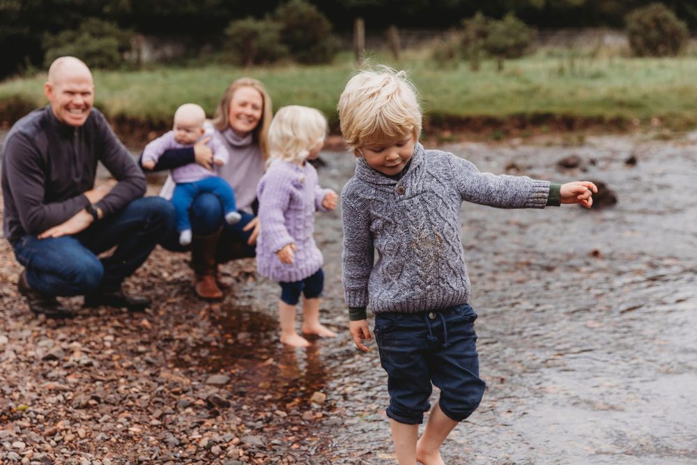 Family enjoying a day by the water, with children playing on a rocky shore while parents smile and watch.