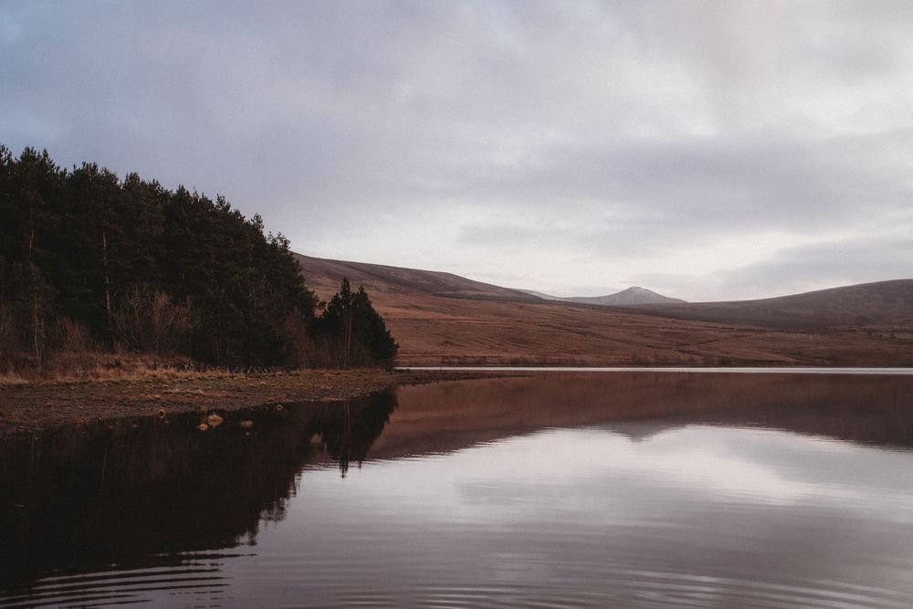 Calm lake reflecting trees and a mountain peak under overcast skies in a serene highland landscape.