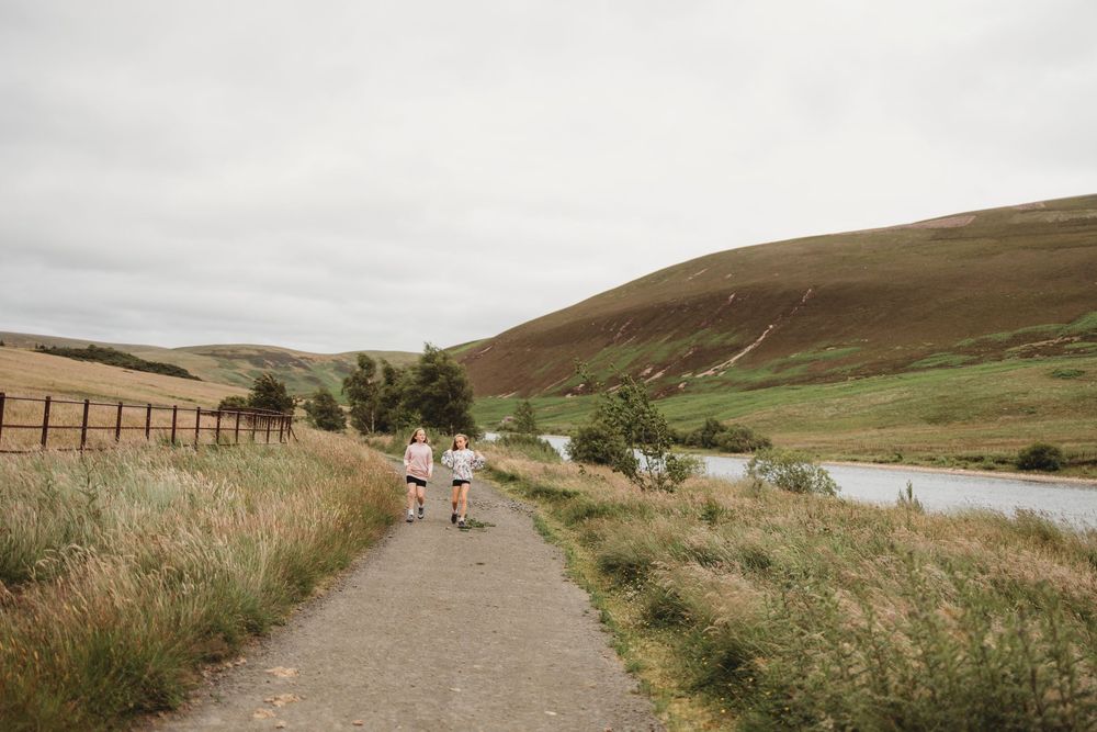 Two people walking on a scenic path beside a calm lake with rolling green hills under an overcast sky.
