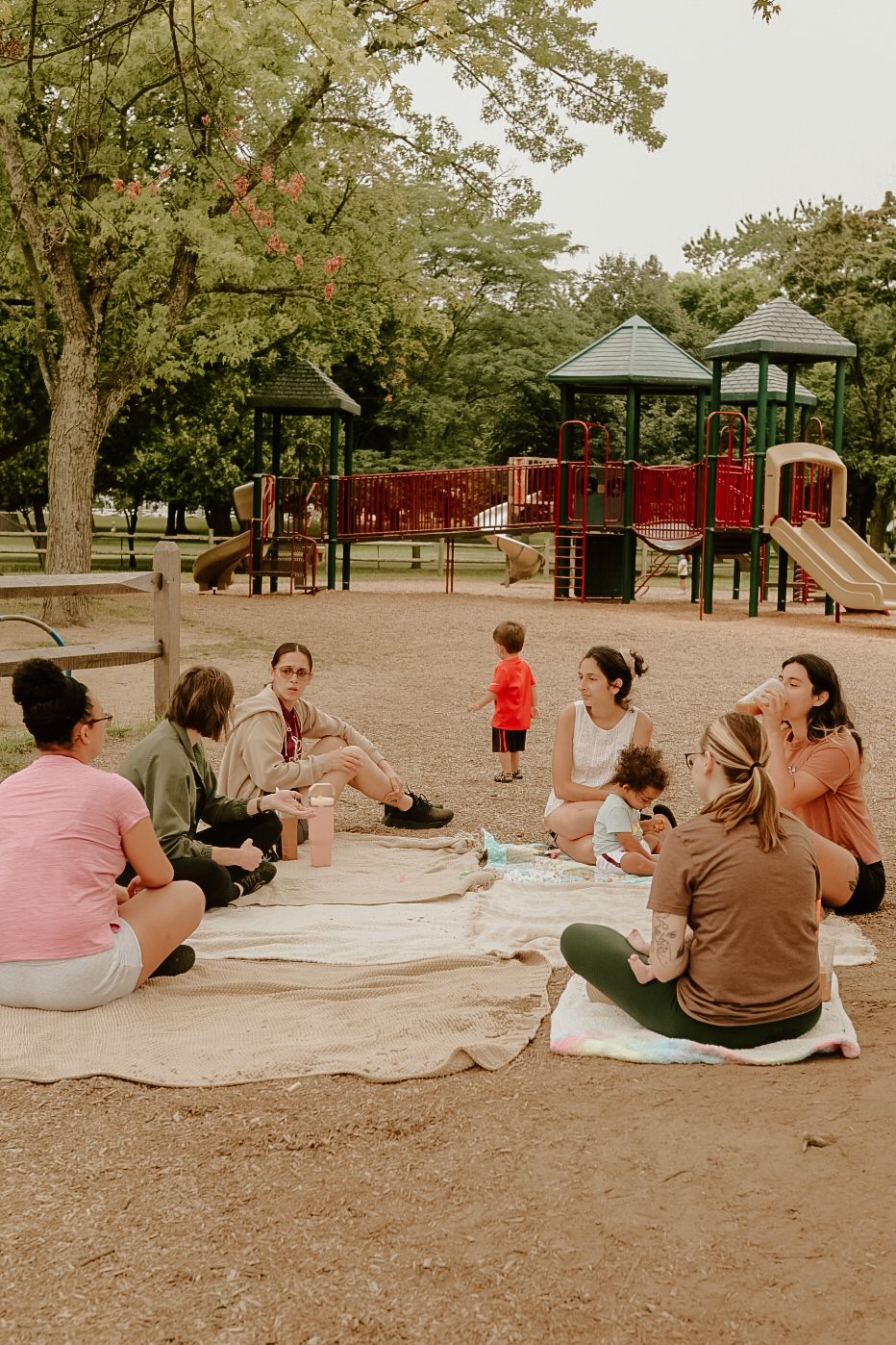 Group of mothers sitting on blankets at a park playground for an outdoor playdate gathering.