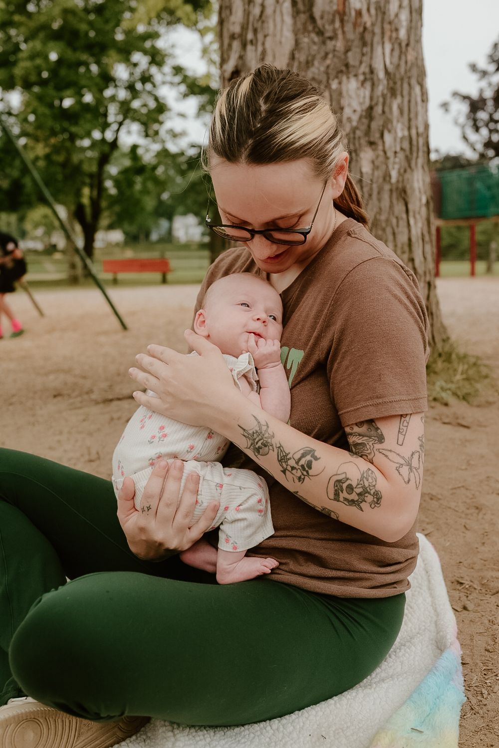 Tattooed mother tenderly holding newborn baby while sitting outdoors in a park near a tree.
