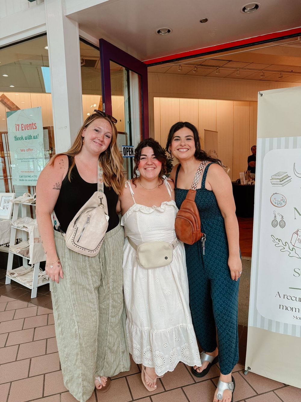 Three women smiling together outside a boutique shop, wearing summer outfits with crossbody bags.