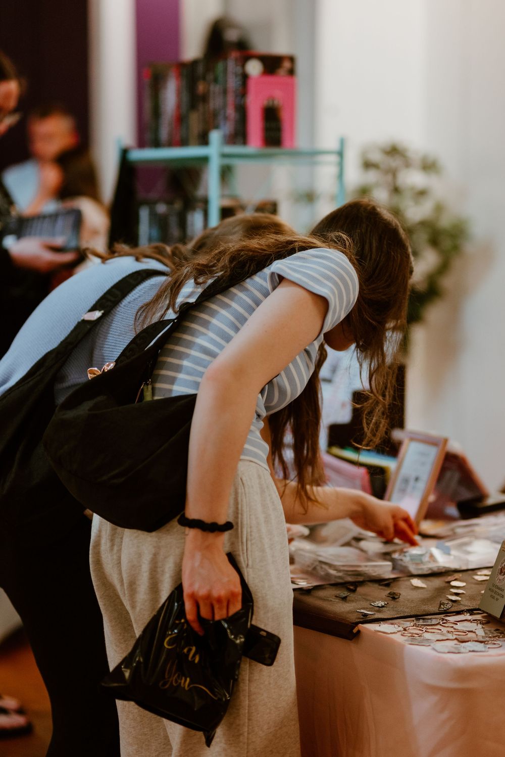 Woman with backpack leaning over a table examining small items at an indoor market or craft fair.