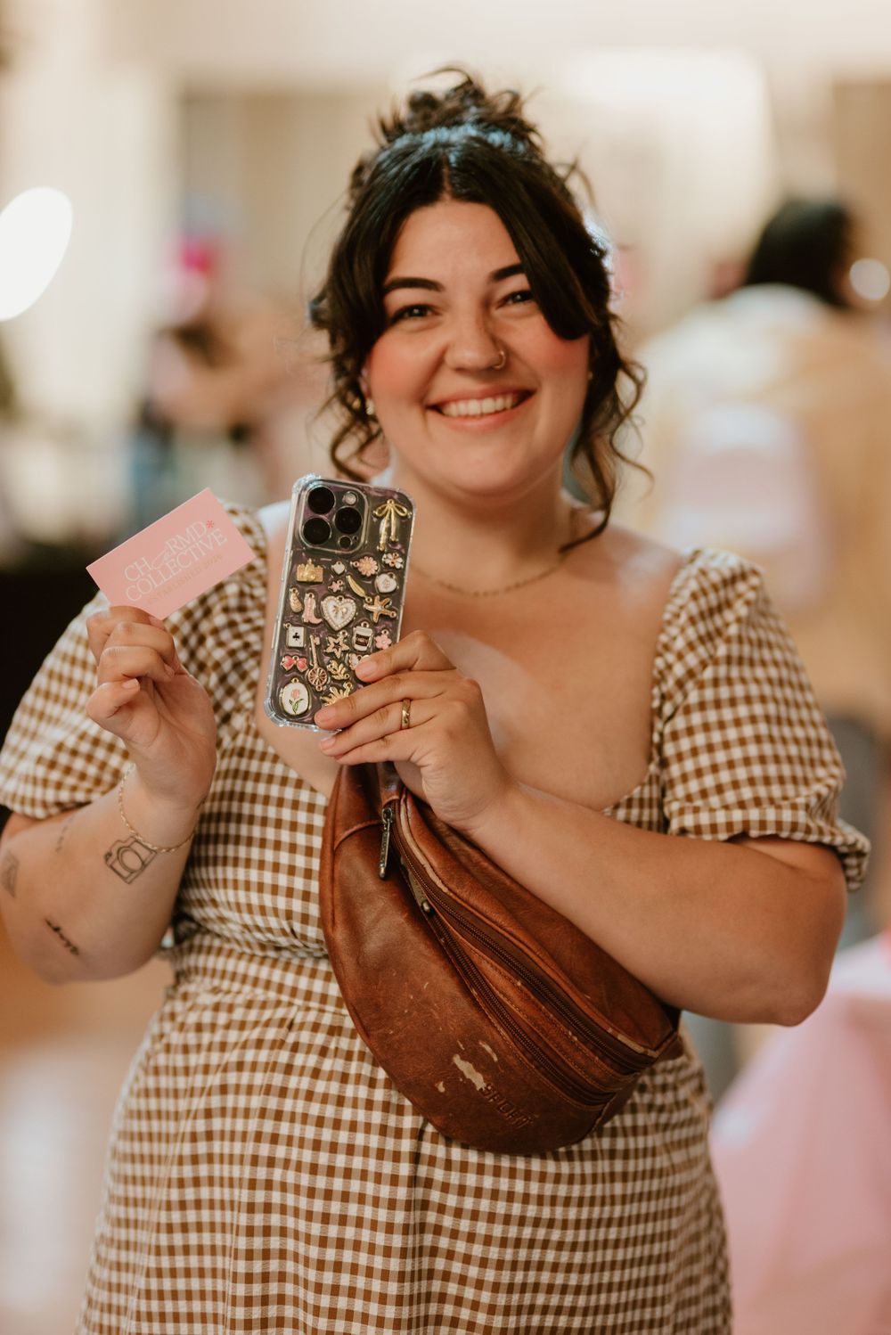 Smiling woman in gingham dress holding decorated phone case and pink card, wearing brown leather belt bag.