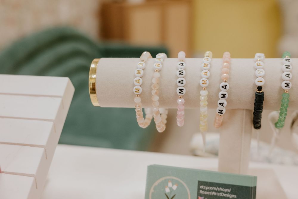 Beaded bracelets spelling MAMA displayed on a jewelry stand, featuring pastel and white beads.