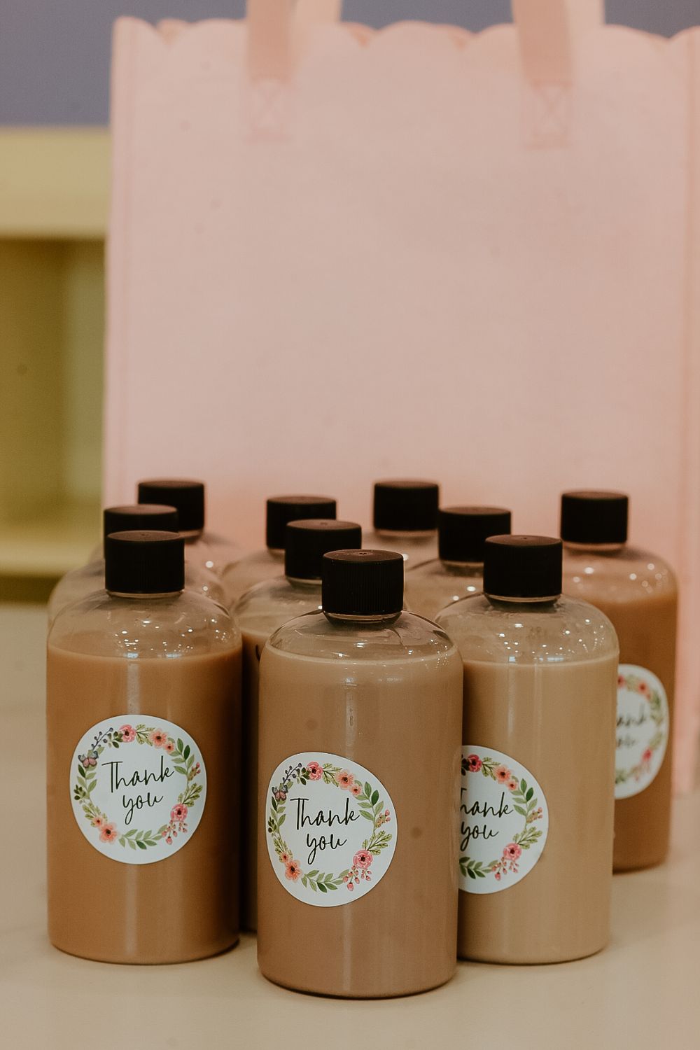 Clear bottles filled with brown liquid, topped with black caps and Thank You floral sticker labels, arranged near a pink gift bag.