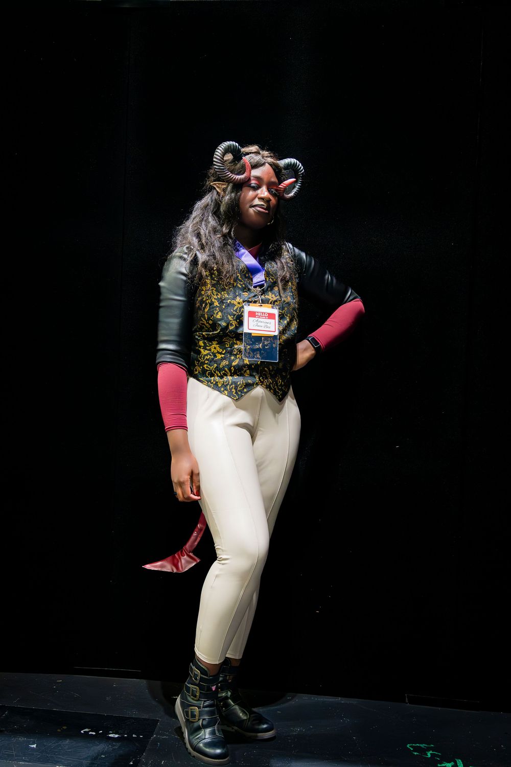 Person in fantasy cosplay costume with horns, ornate vest, white pants, and boots poses against black background.