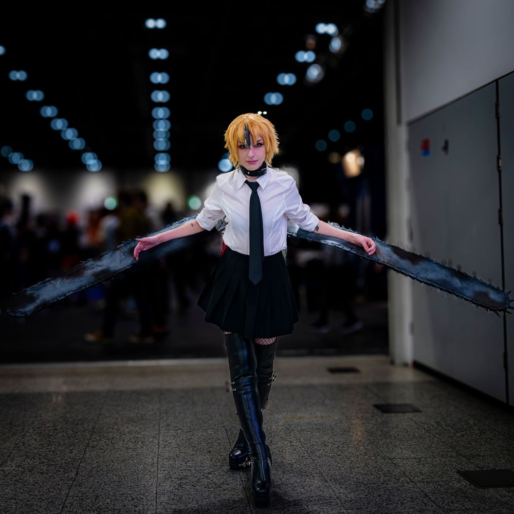 Cosplayer in school uniform with black wings poses dramatically at anime convention.