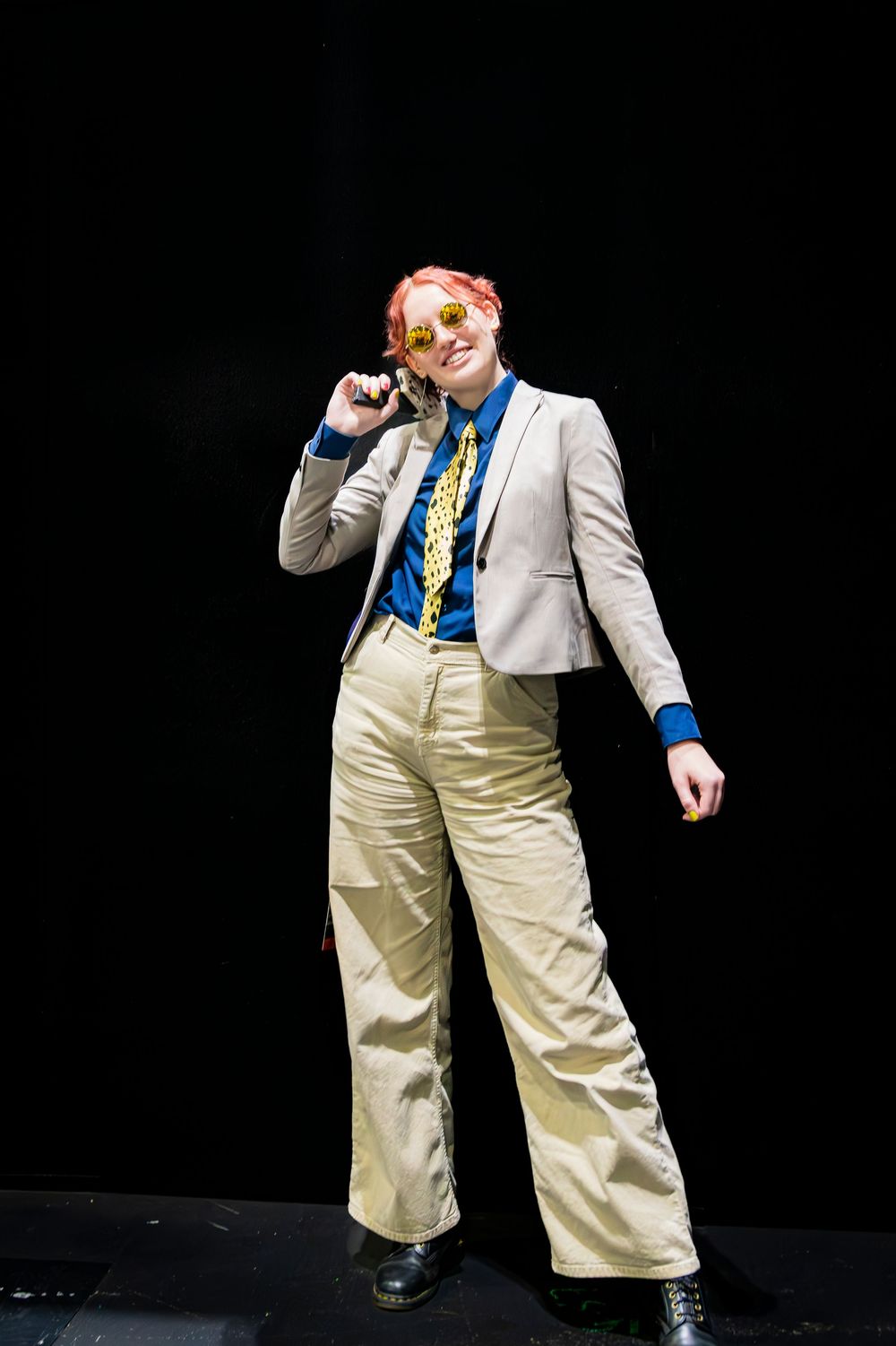 Person in beige suit with pink hair and gold glasses poses on dark stage.