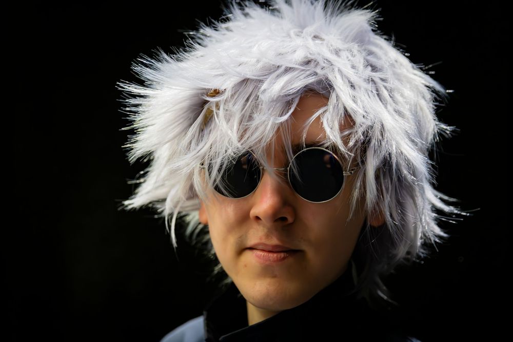 Person wearing a wild white wig and round sunglasses cosplaying against a dark background.