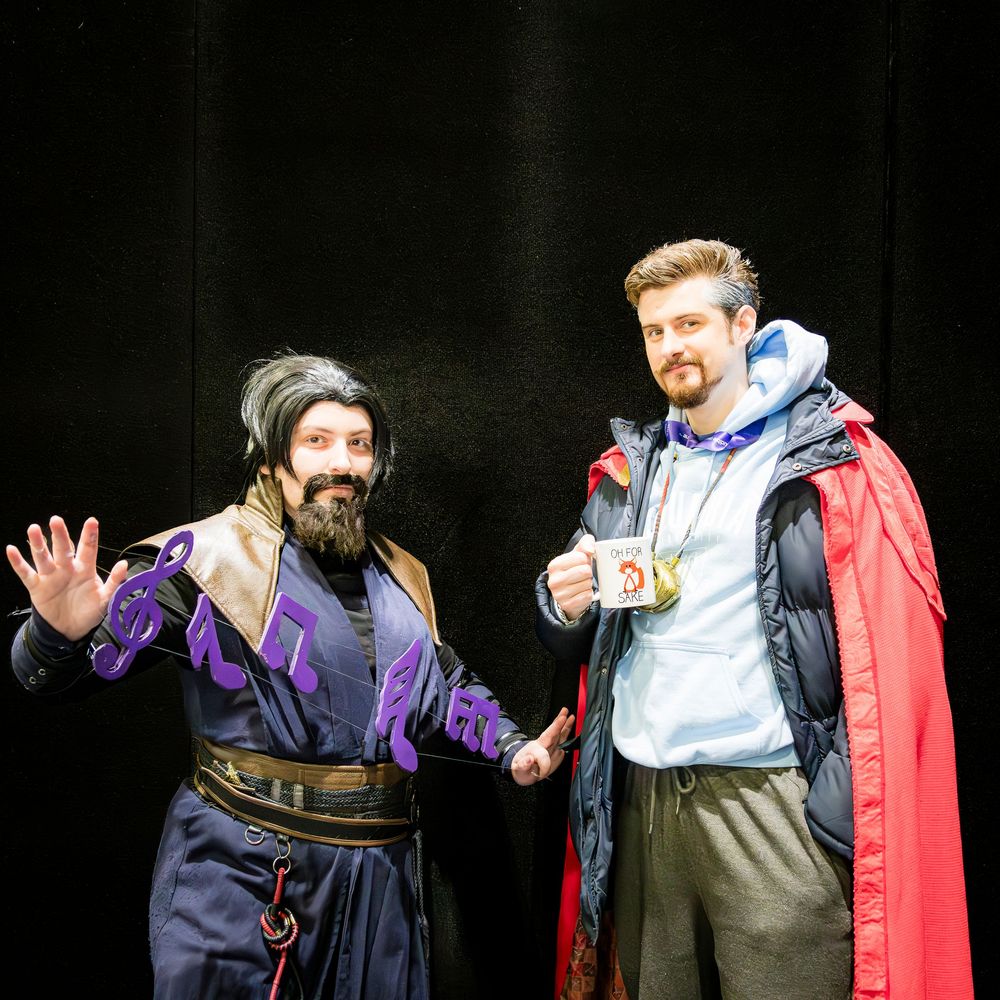 Two cosplayers in fantasy costumes smile and pose against black background.