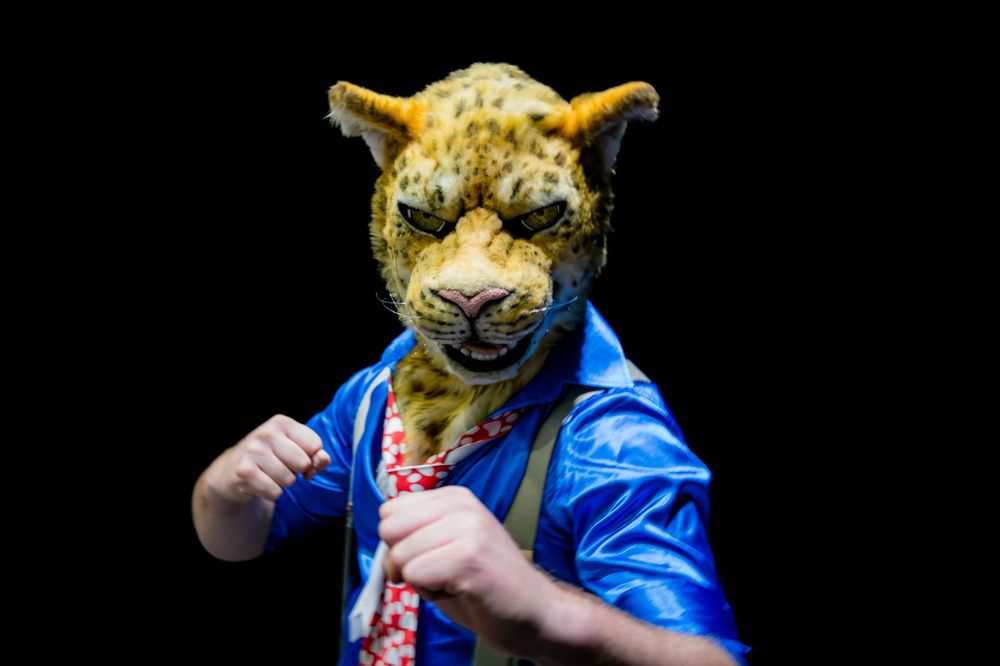 Person wearing a realistic leopard mask and blue jacket with a red tie poses against a black background.