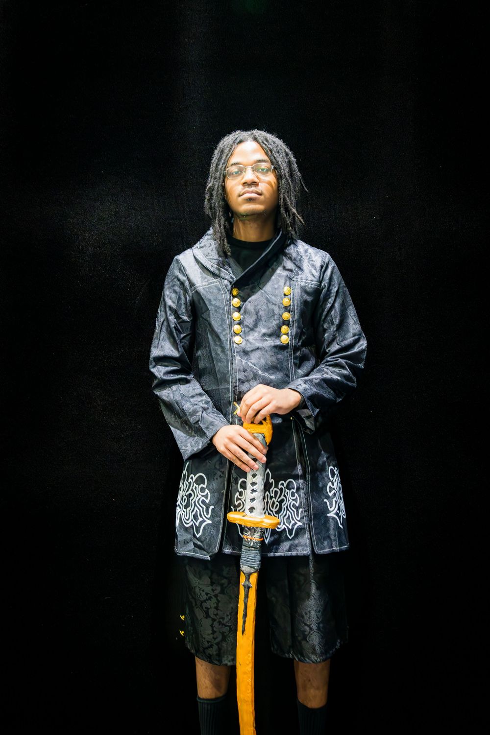 Person in detailed dark samurai costume holding a katana with yellow handle against black backdrop.