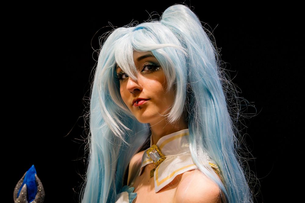 Woman in blue anime cosplay wig and costume posing against dark background.