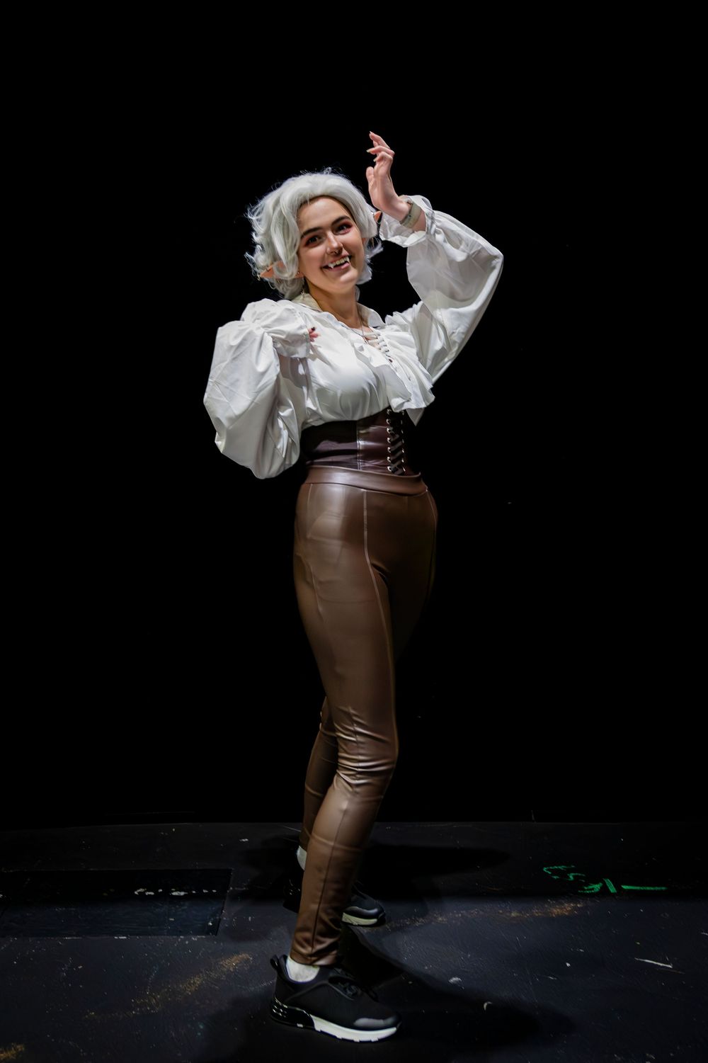 Woman in white ruffled blouse, brown corset, leather pants, and white wig poses on dark stage.