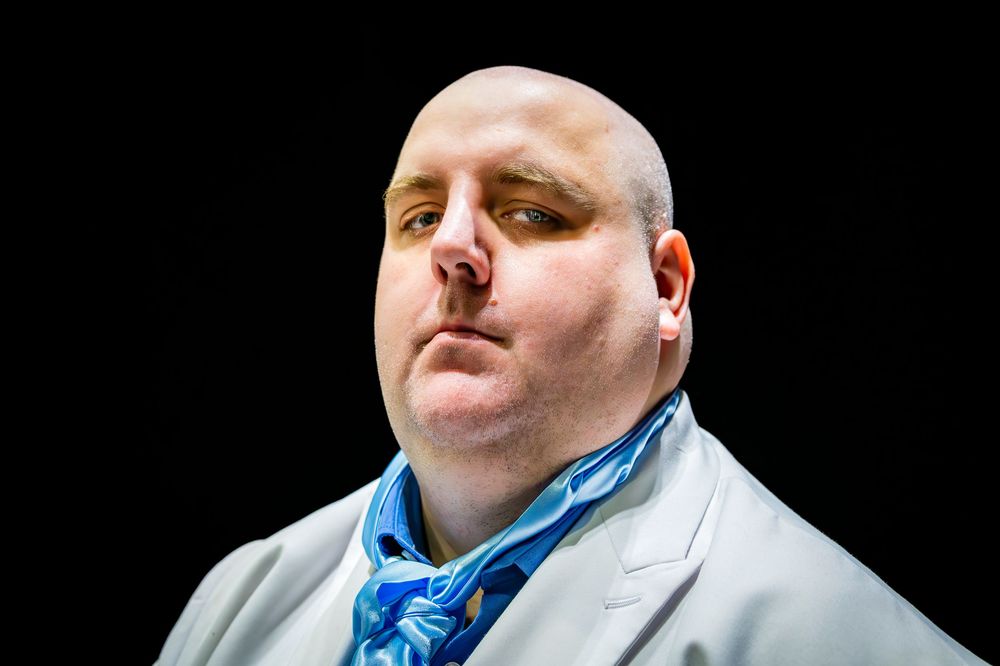 Bald man in gray suit with blue tie posing against dark background.