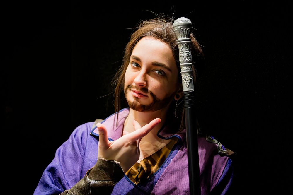 Person in purple costume with beard making hand gesture, holding decorative staff, theatrical performance portrait.