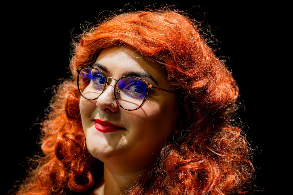 Woman with curly red hair and blue glasses smiling against a dark background.