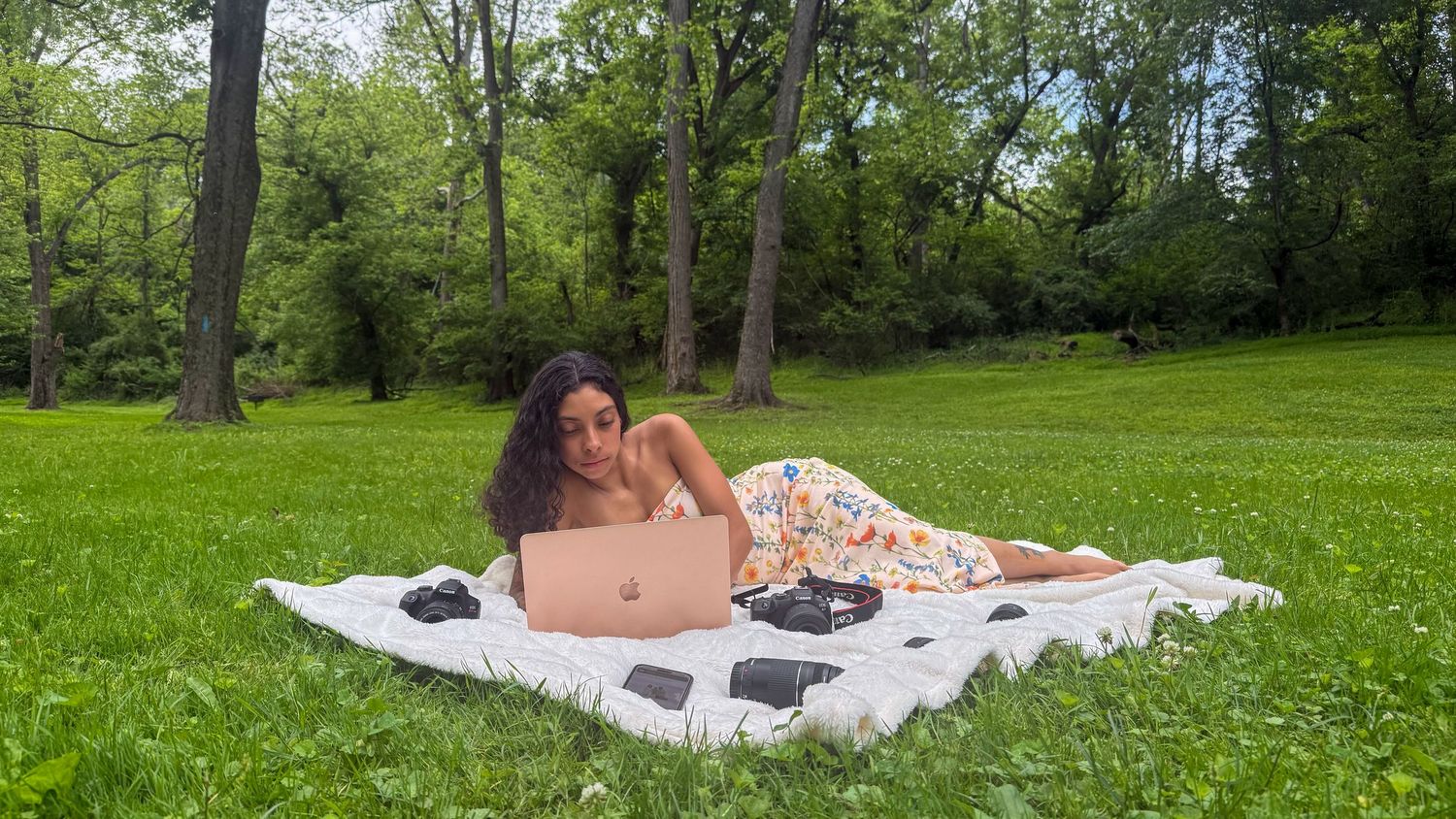A young woman lies on a blanket in a lush green park, working on a laptop on a sunny day.