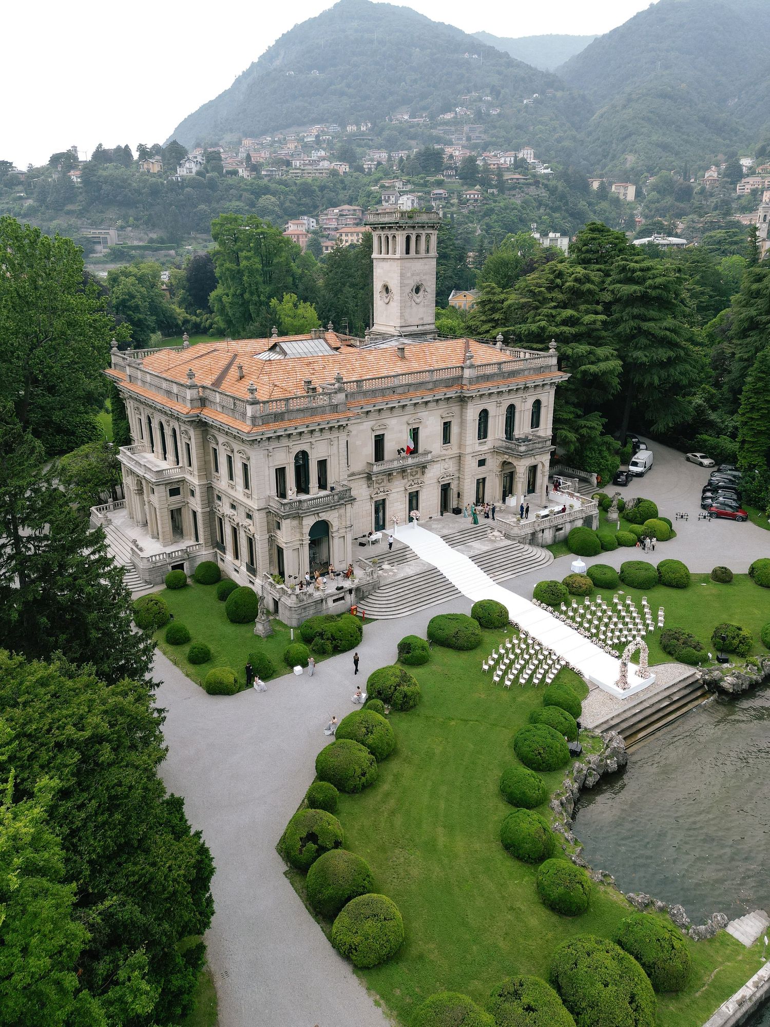 Aerial view of an elegant Italian villa with manicured gardens and curved driveway overlooking Lake Como and mountains.