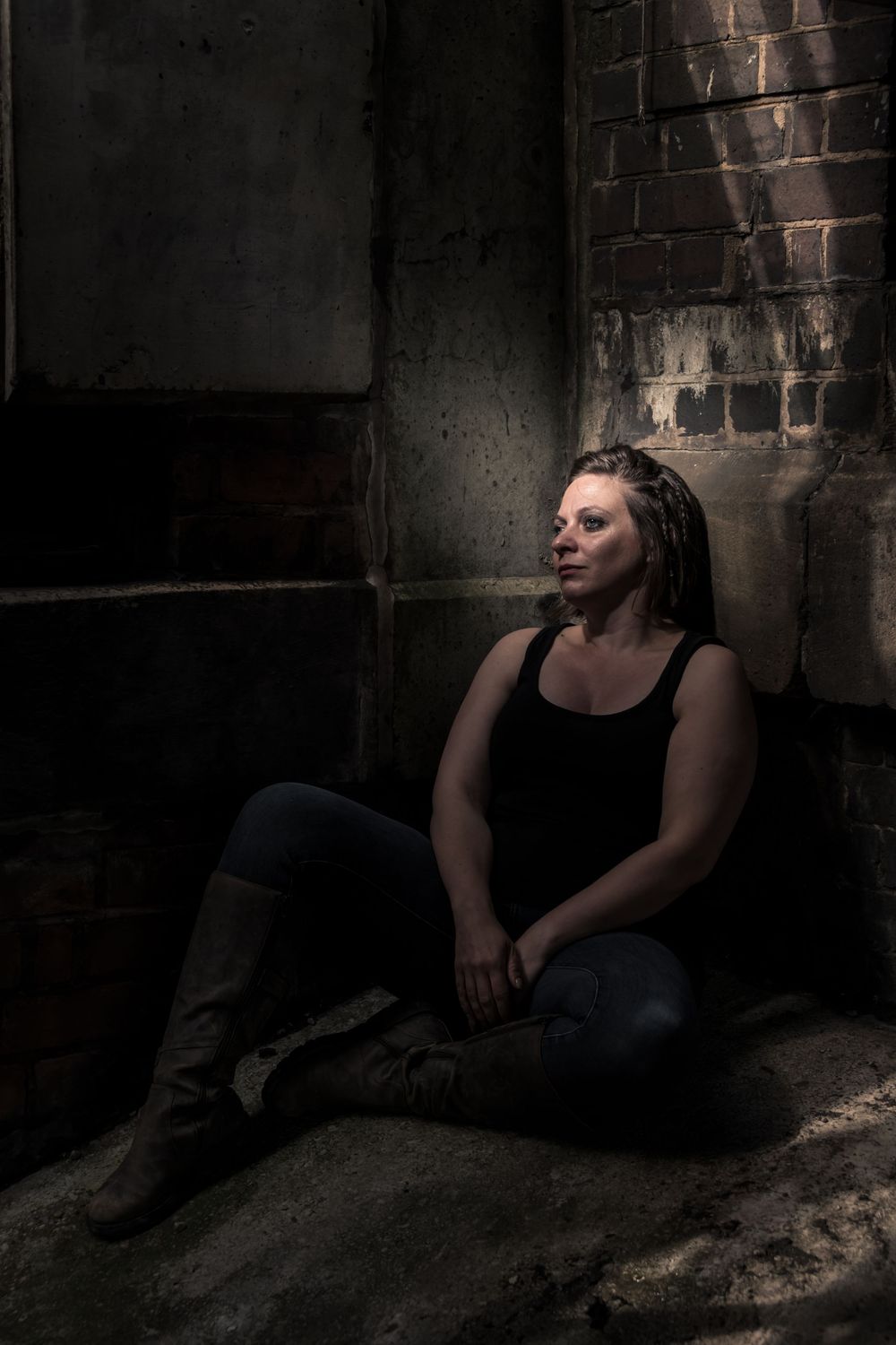 Woman with braids sits against dark brick wall in moody lighting, wearing black tank top, jeans, and knee-high boots.