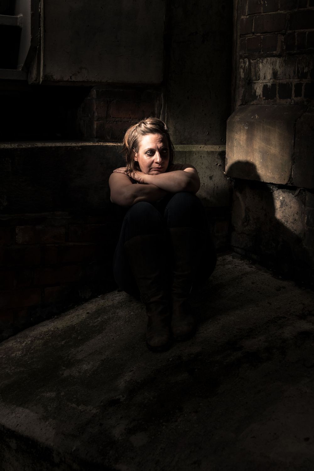 A distressed woman sitting alone in a dark, abandoned building, hugging her knees in a somber pose.