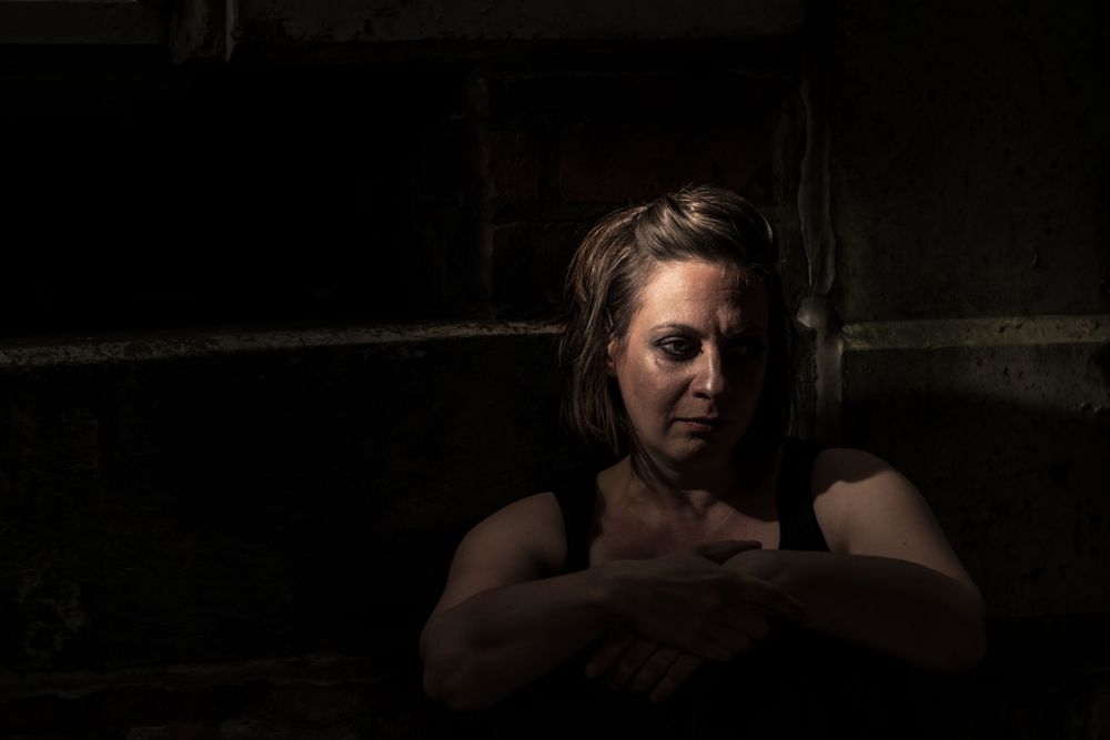 A woman sits alone in dim light, arms crossed, with a somber expression in a dark, moody setting.