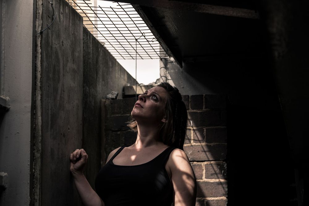 Woman in black tank top leaning against brick wall in dramatic dark alleyway with light streaming through grate above.