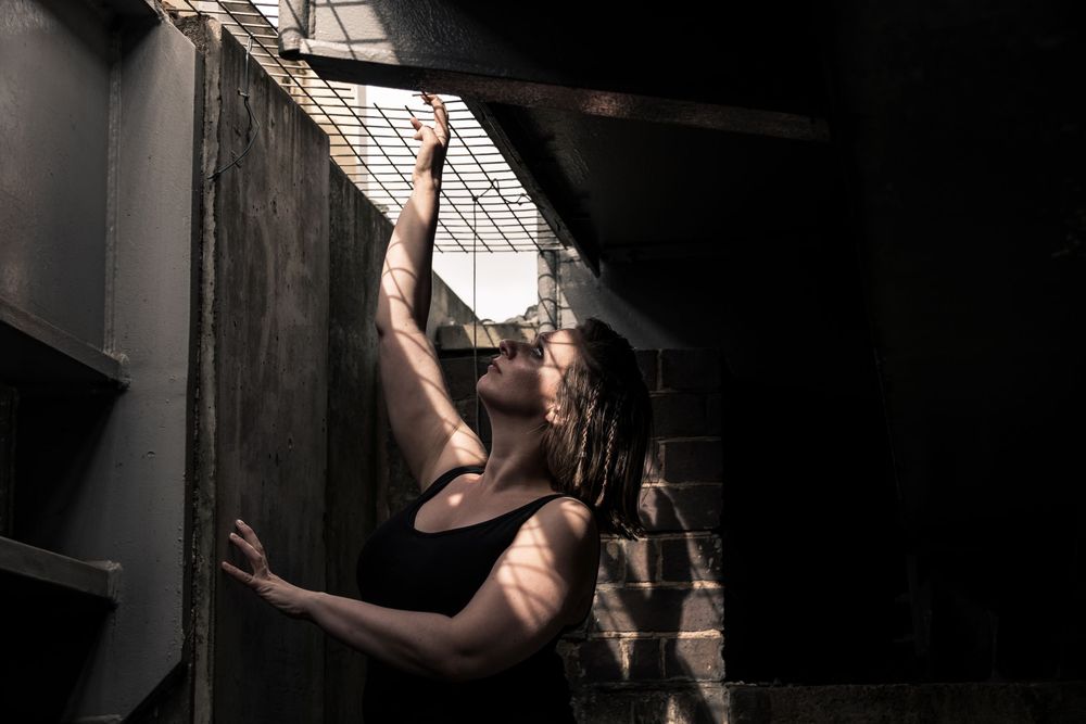 A person in a black tank top leans against a dark industrial wall, reaching upward toward a metal grate window.