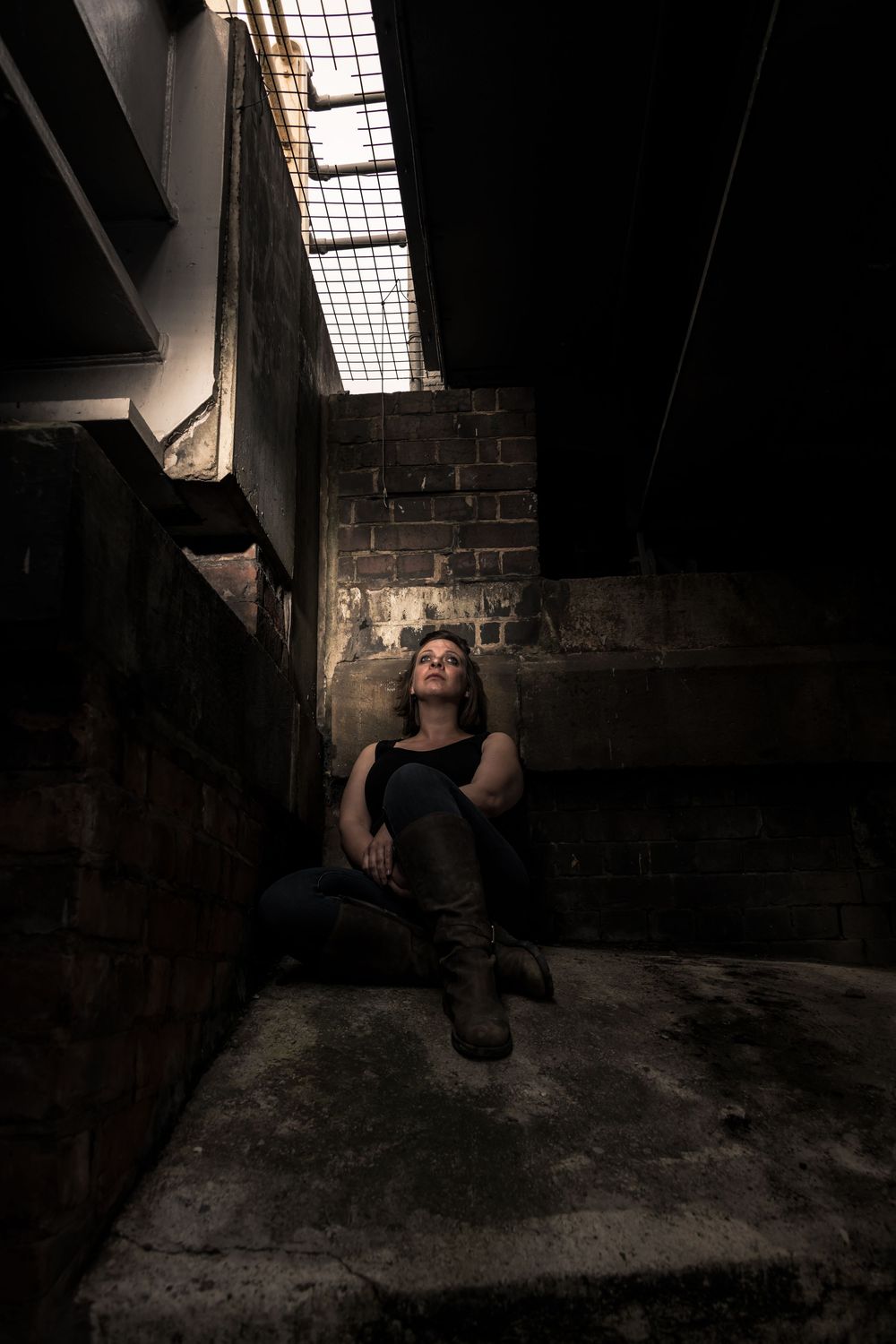 Woman in black tank top and boots sitting in a dark abandoned building looking up at a barred skylight.