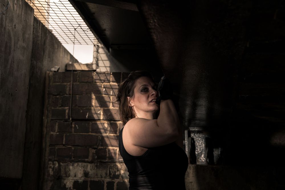 Woman in black top leaning against concrete pillar in dramatic shadowy industrial urban setting.