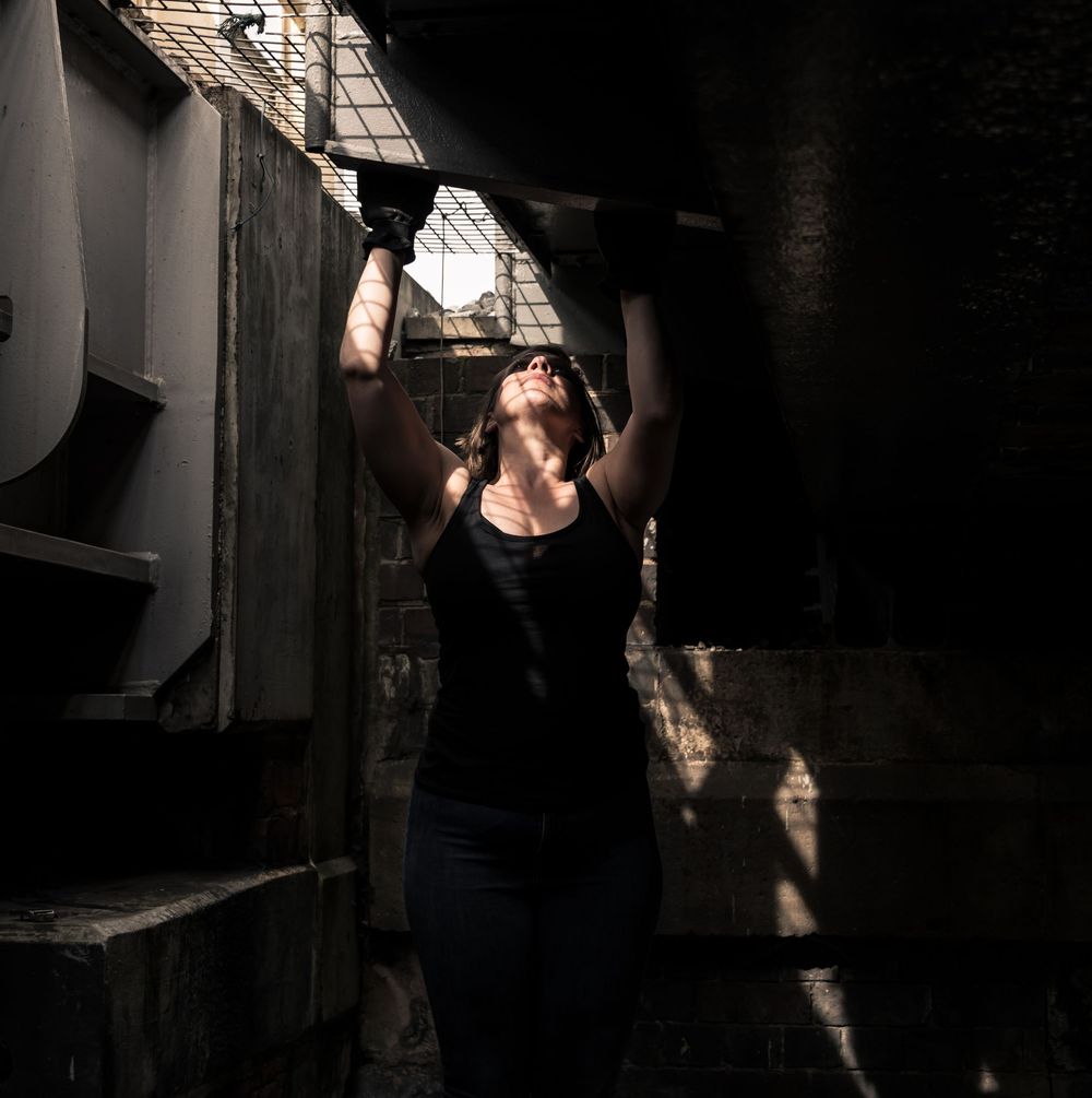 Woman in black tank top reaches upward in dramatic industrial setting with striking light and shadow contrast.