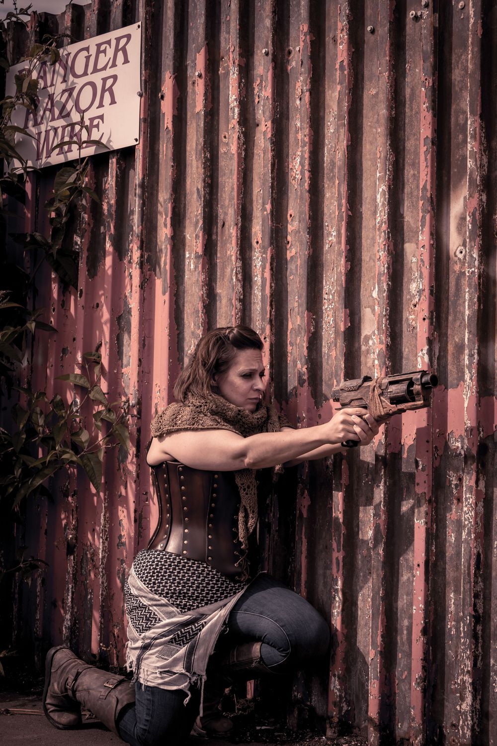 A woman in a corset and tactical gear aims a pistol while kneeling against a rusty corrugated metal wall outdoors.