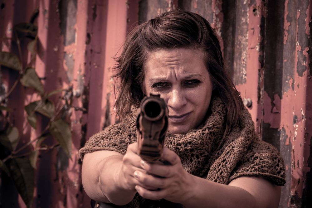 Woman aiming a rusty pistol with intense focus, standing against a weathered red metal wall outdoors.