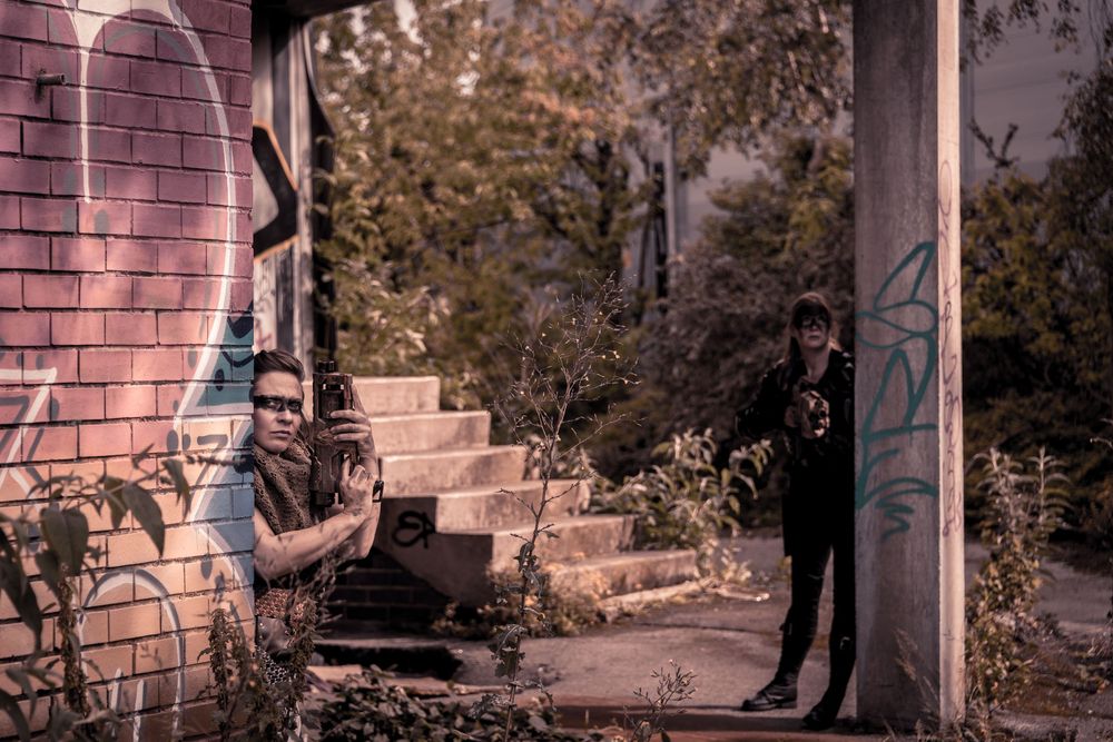 Two people pose in an urban abandoned building with graffiti-covered walls and overgrown vegetation.
