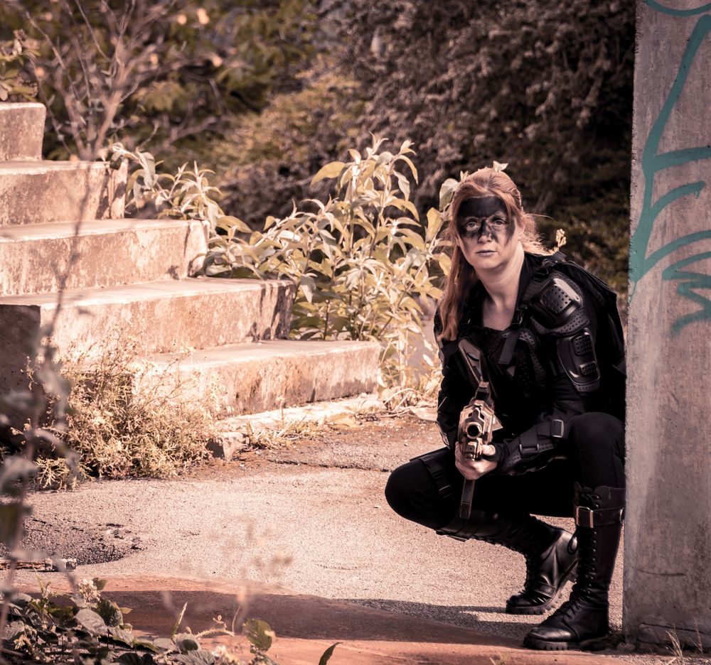 Woman in black leather costume and mask crouching outdoors near graffiti wall and stone steps.