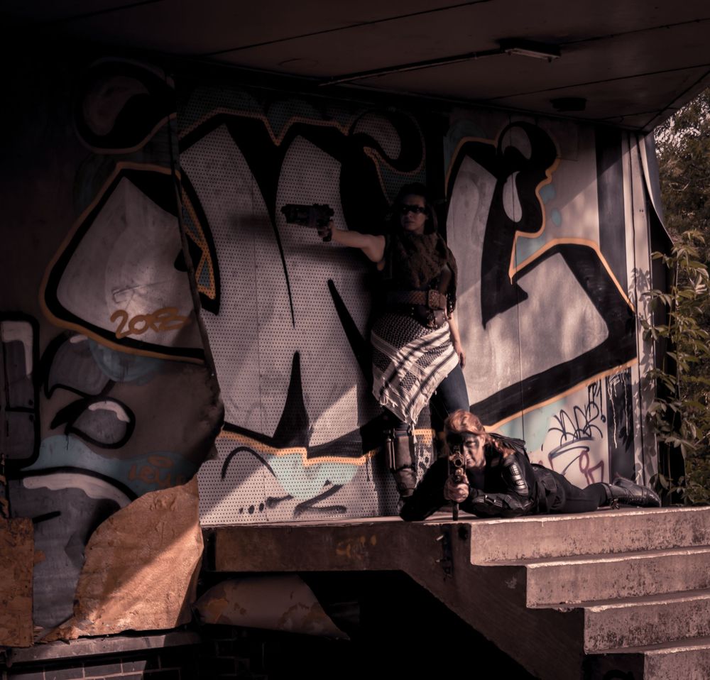 Two people pose near graffiti-covered walls under a bridge in a dark, urban setting.