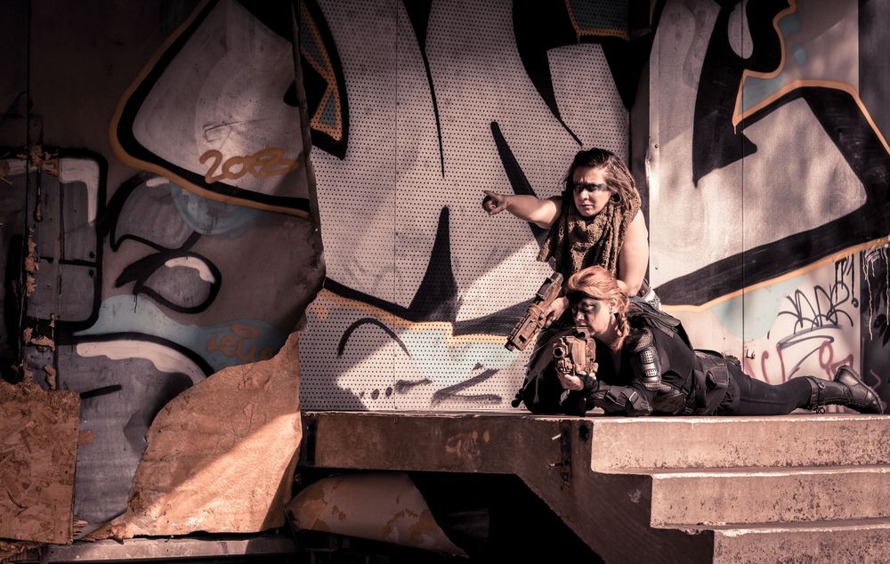 A wild-looking figure with long hair crouches on concrete steps before a colorful graffiti wall in an urban setting.