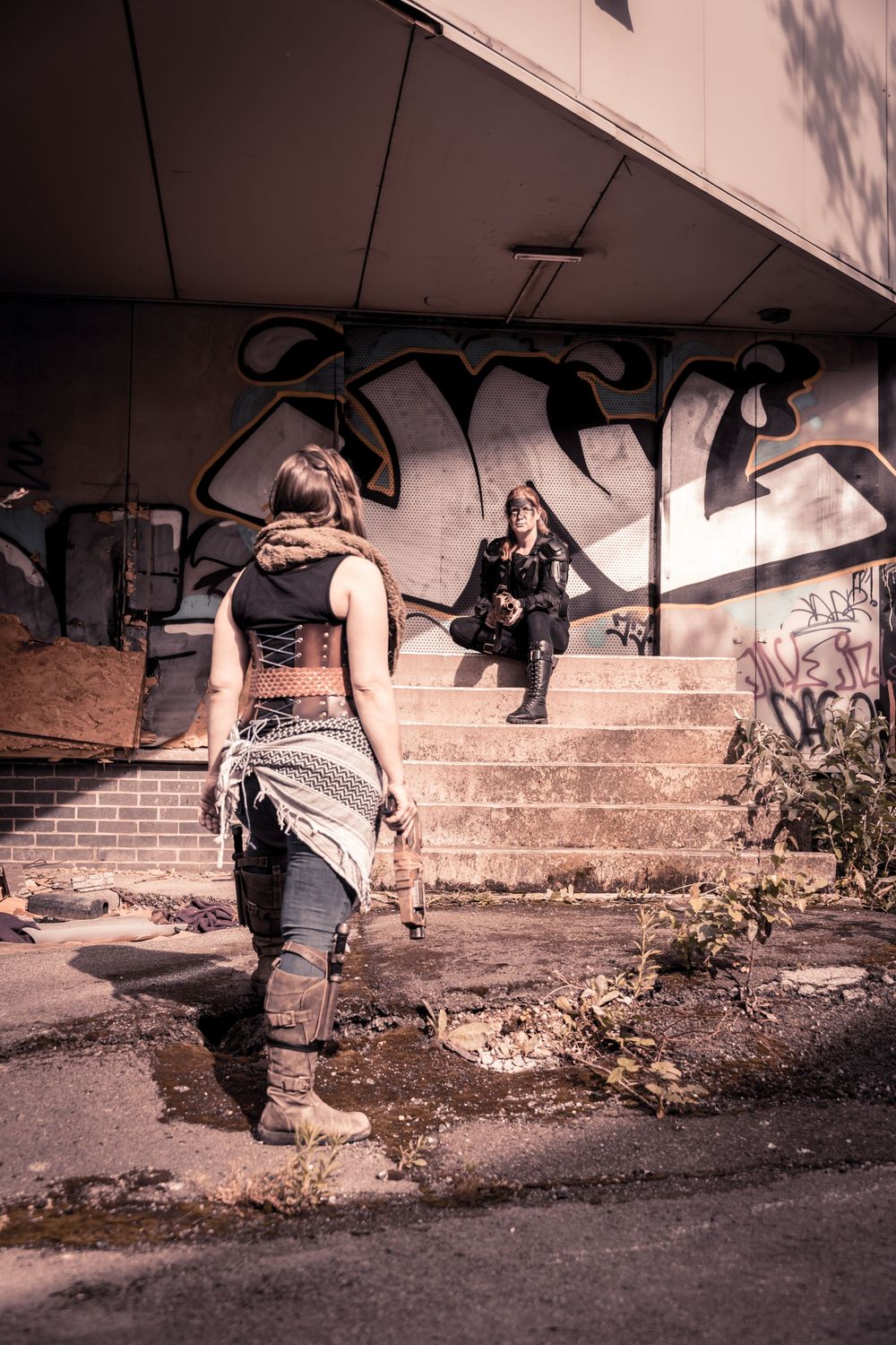 Two women in post-apocalyptic outfits near graffiti-covered steps of an abandoned urban building.