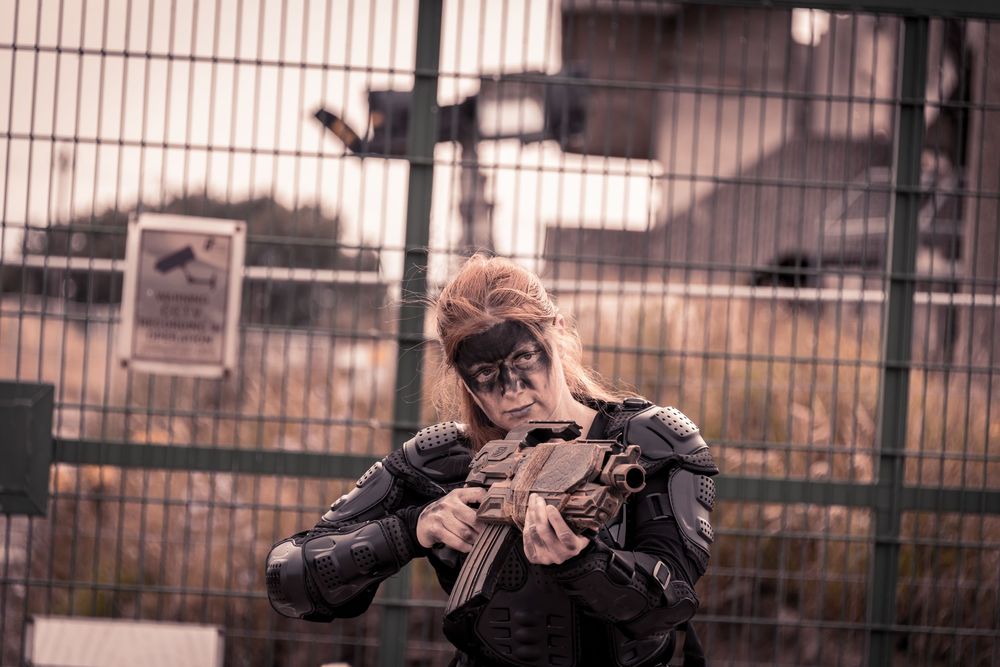 Woman in tactical armor and face paint aims a rifle near a chain-link fence in a post-apocalyptic cosplay scene.