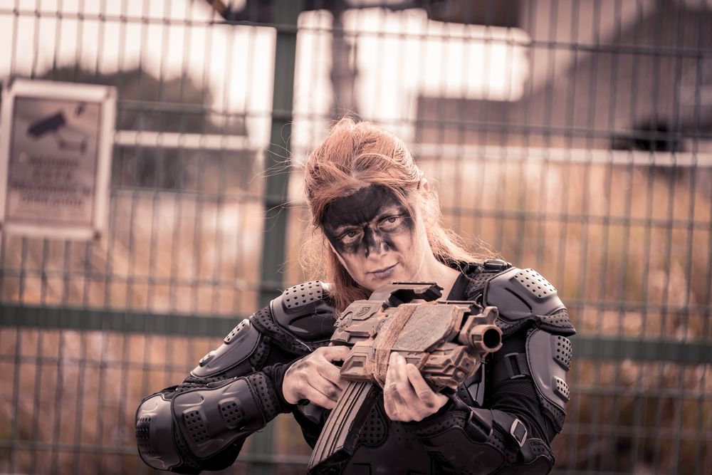 Woman in tactical armor and face paint holds assault rifle in post-apocalyptic cosplay near chain-link fence.