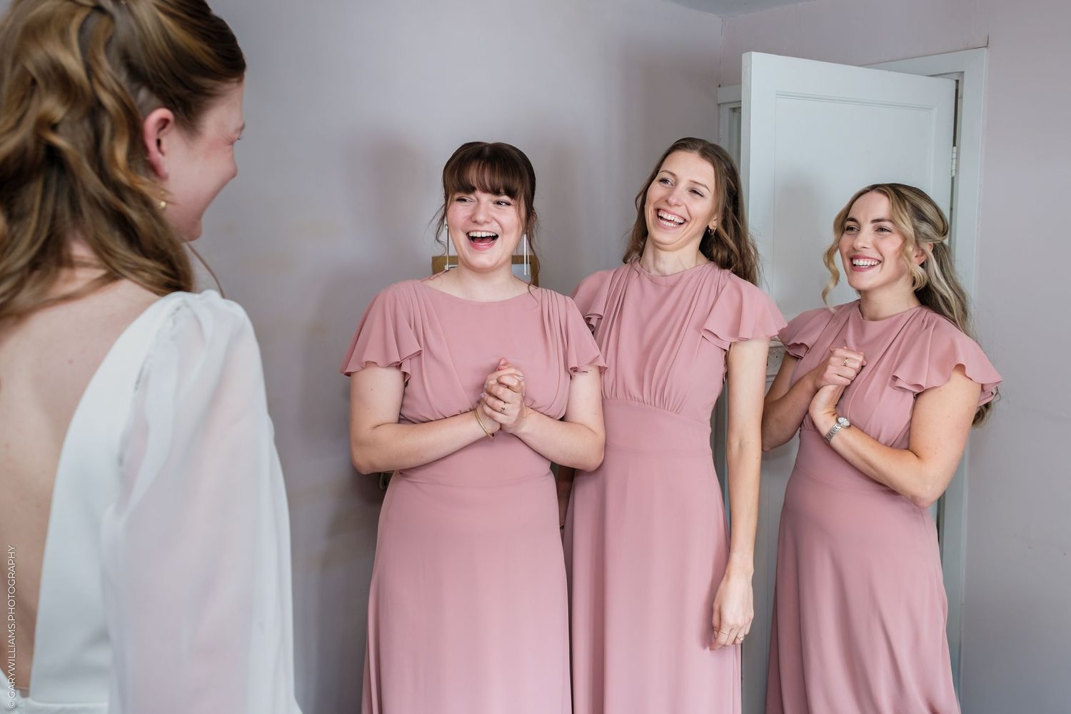 Three bridesmaids in pink dresses react with joy and smiles while seeing the bride in her wedding gown.