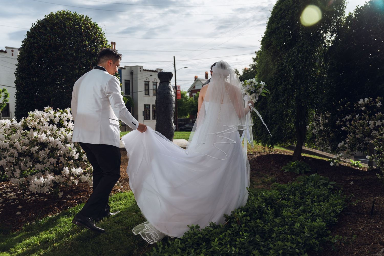 Groom helps bride with wedding dress train in a lush garden with blooming flowers.